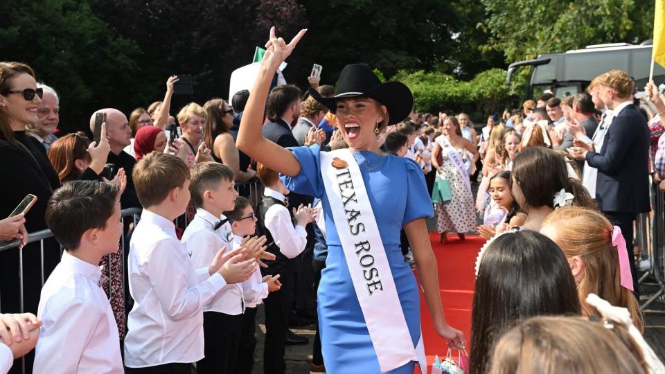 Woman wearing a Texas Rose sash and cowboy hat making the Hook ’em Horns gesture amid a cheering crowd
